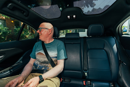Robin Spinks sits in the back seat of a Waymo vehicle, a Jaguar I-PACE, wearing a blue graphic T-shirt, khaki trousers, and blue glasses. He is smiling and looking out the window while sunlight filters through the panoramic glass roof of the car. He is wearing a seatbelt.