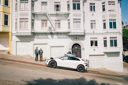 Newton and Steve standing on a sloped sidewalk in front of a white building, as a white Waymo autonomous vehicle pulls up in front of them