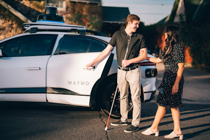Lilian holding onto Max's arm as he opens the door to a Waymo One autonomous vehicle