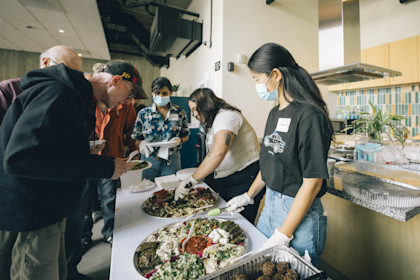 Volunteers stand in a kitchen, serving Mediterranean food to a line of older men